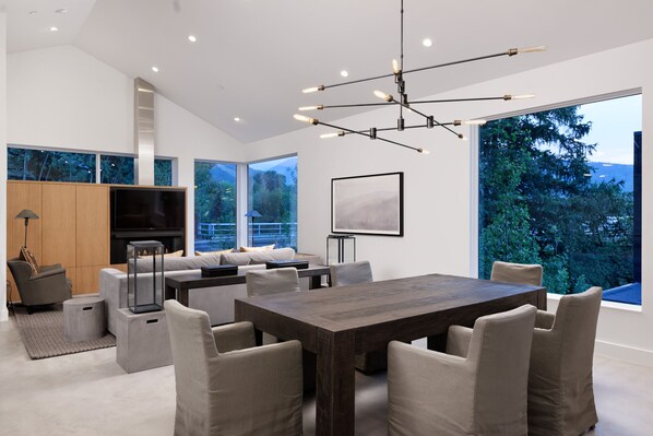 A modern living room with a large dining table, comfortable seating, a fireplace, and large windows offering a view of the outdoors.