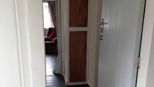 Un couloir étroit avec une porte en bois et un sol recouvert de moquette, qui mène à une pièce où se trouvent un fauteuil rouge et des rideaux.