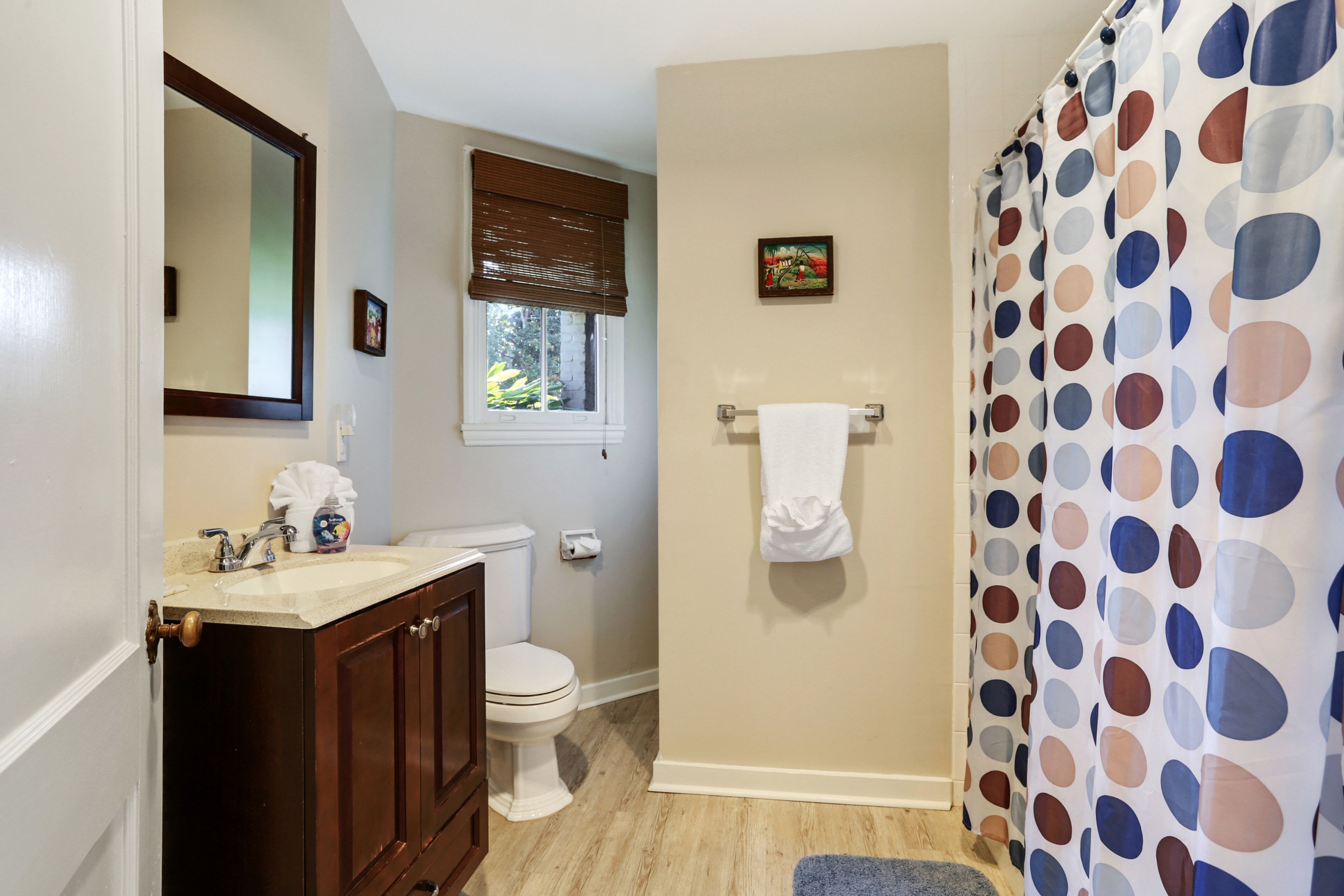 Une salle de bain avec un meuble-lavabo en bois, des toilettes, une fenêtre avec des stores, un porte-serviettes et un rideau de douche à motifs à pois.