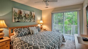 A bedroom with a bed, a ceiling fan, a nightstand with a lamp, a window with blinds, and a view of greenery outside. - 5 Mi to Disney: Emerald Island Resort Condo! (Kissimmee)