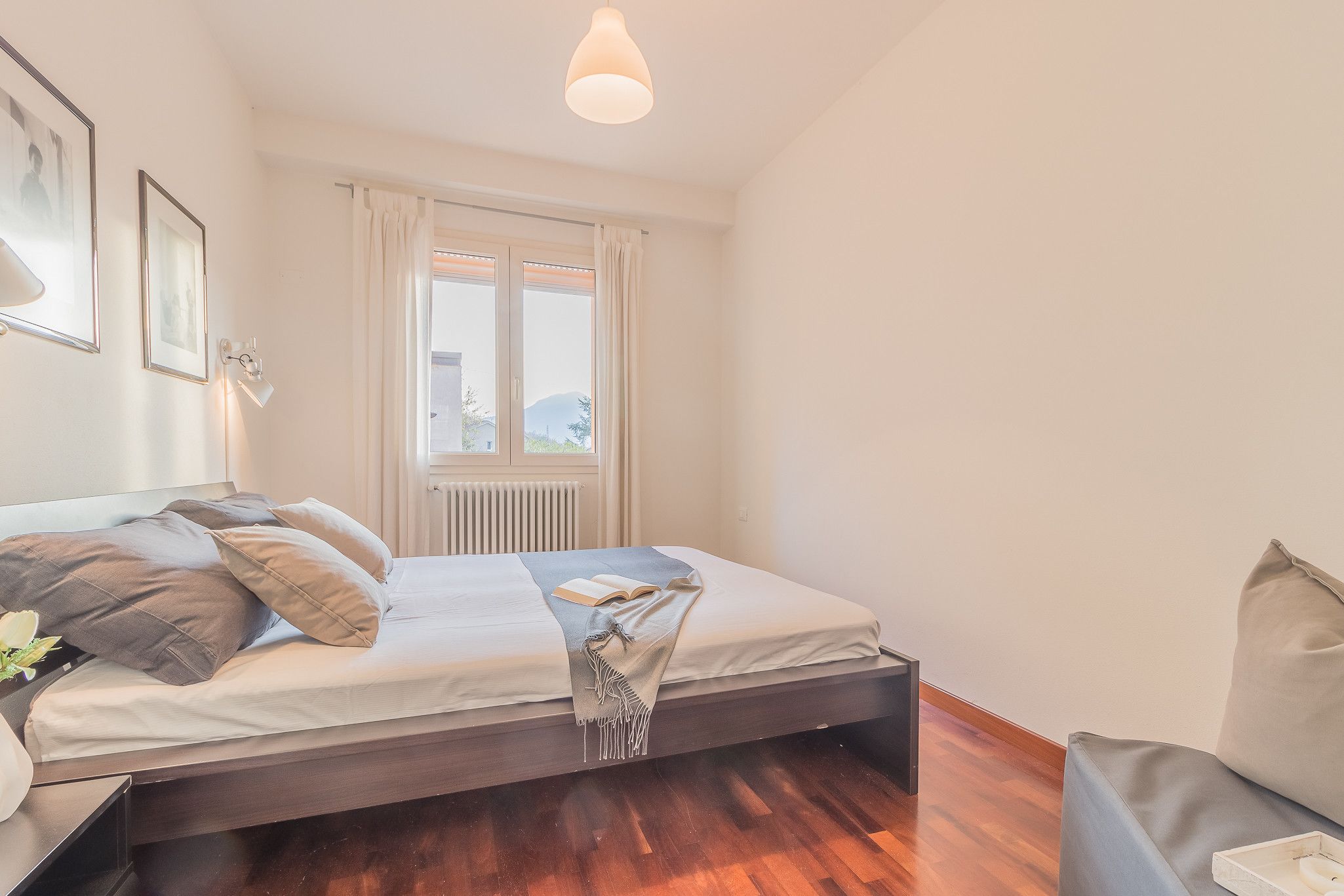 A bedroom with a bed, a bedside table, a window with curtains, and a view of a building outside.
