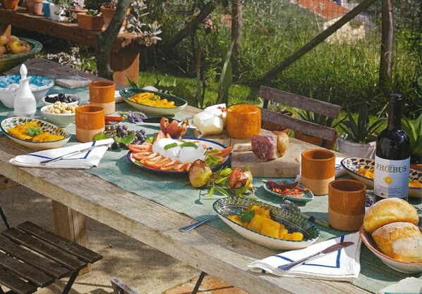 A wooden table set with various dishes, including fruits, cheeses, and bread, outdoors with greenery in the background.