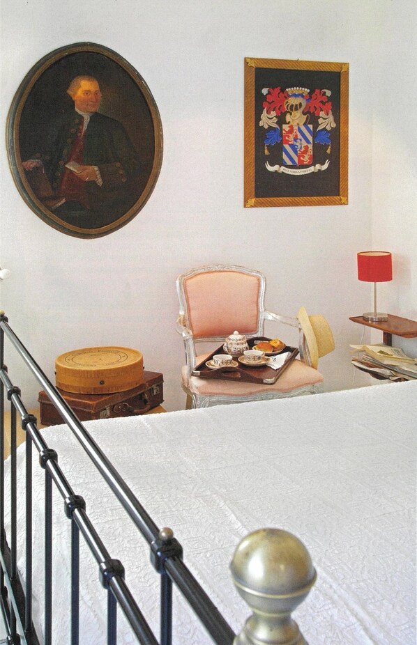 A room with a bed, a chair with a tray of tea and pastries, a painting, and a coat of arms on the wall.
