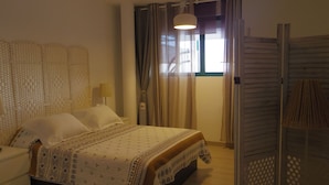 Un dormitorio ordenado con cama, mesitas de noche, lámpara, ventana con cortinas y un radiador fijado a la pared.
