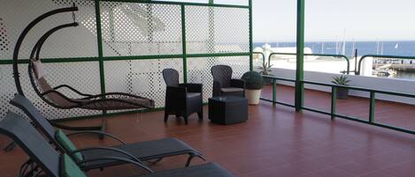 Un área de estar al aire libre con techo, sillas y mesas, y vista al mar, con piso de baldosas.