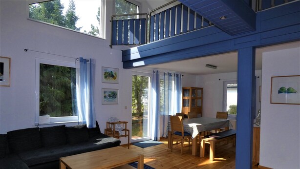 Ett rymligt vardagsrum med en soffa, ett träbord och ett matbord med stolar. Det finns en balkong med ett räcke och utsikt över träd.