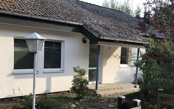 A white house with a tiled roof, two windows, a small porch, and a garden area with rocks and plants. - Large house with lake access (Zehdenick)