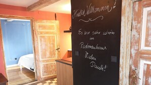 A room with a checkered floor, a bed, a wooden door, and a chalkboard with text.