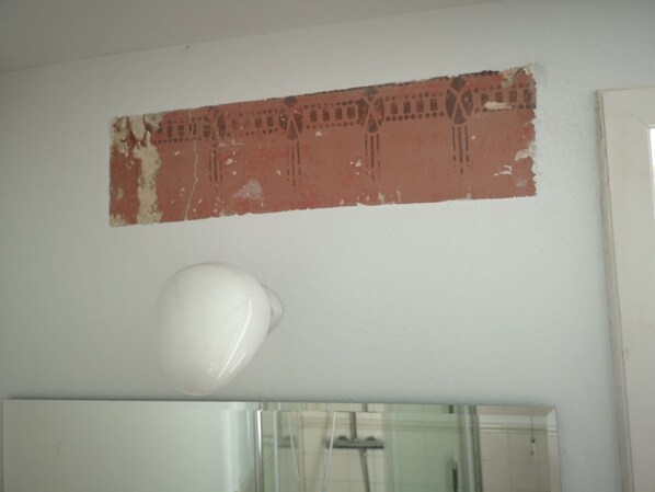A bathroom with a white wall, a partially damaged red decorative panel, and a white floor lamp. - Holiday in historic thatched-roof estate from 1750 (Norderbrarup)