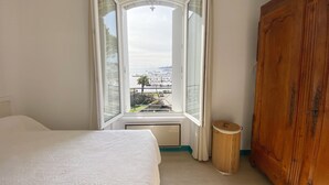 Une chambre avec une grande fenêtre, une armoire en bois et un lit recouvert d’une courtepointe blanche.