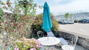 Une terrasse avec une table et des chaises, un pot de fleurs orné de fleurs roses, un parasol bleu et une vue sur une marina avec des bateaux.