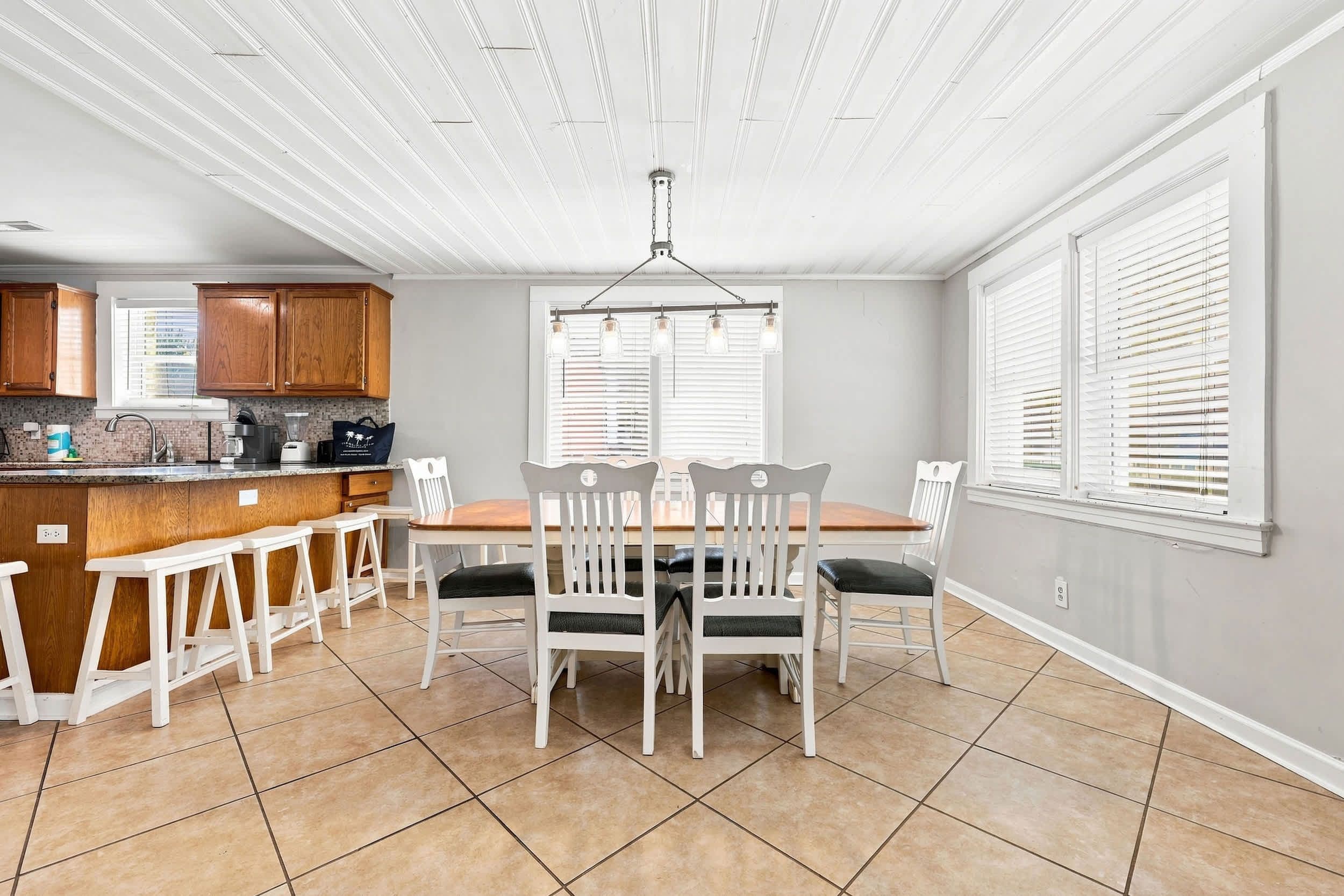 A kitchen with a dining area, featuring a central island, white chairs, and a hanging light fixture.