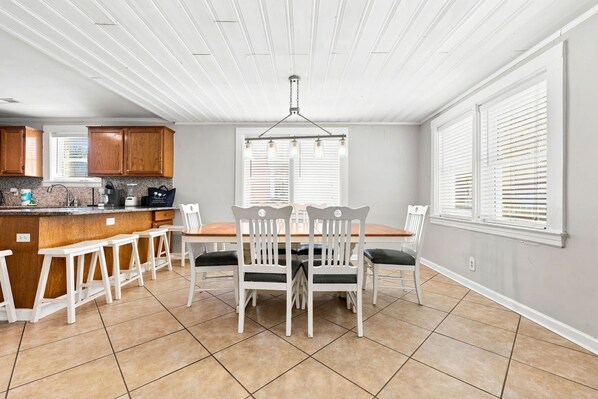 A kitchen with a dining area, featuring a central island, white chairs, and a hanging light fixture.