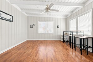 Una habitación con piso de madera, ventilador de techo, ventana con persianas y una mesa con sillas.