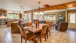 A spacious kitchen with wooden cabinets, a dining area with a wooden table and chairs, a living room with a sofa and TV, and a large window providing natural light.