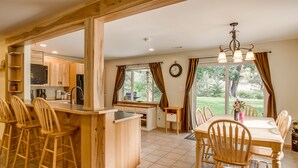 A kitchen with wooden bar stools, a dining table with chairs, and a view of the outdoors through large windows.