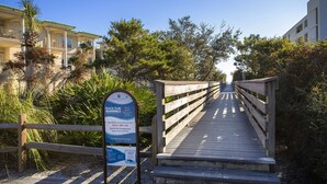 A wooden boardwalk leading to beach access, surrounded by greenery and buildings.