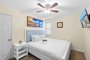 Una camera da letto con un letto, un comodino, una ventola a soffitto, una finestra con persiane e un quadro appeso al muro.