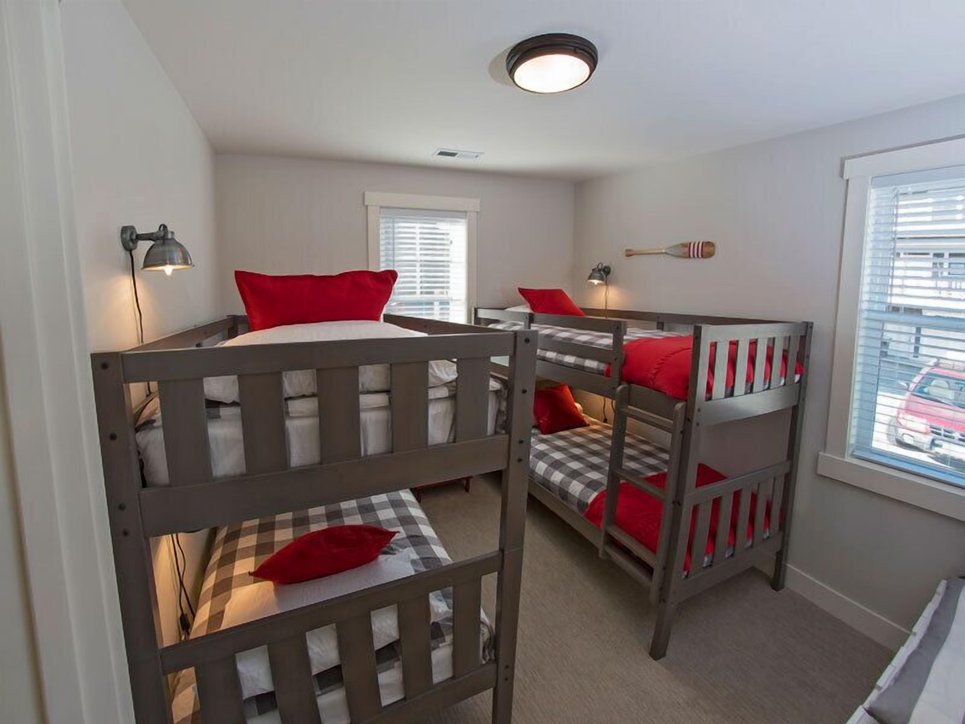 A room with three bunk beds, each with a red pillow and a checkered blanket. There are two wall-mounted lamps and a ceiling light.