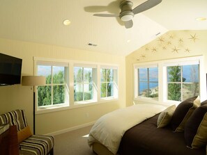 Une chambre à coucher avec un lit, un ventilateur de plafond, une télévision et de grandes fenêtres donnant sur un paysage verdoyant.