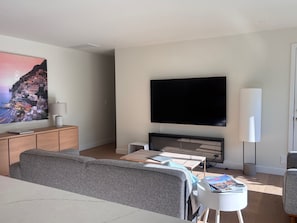 A living room with a sofa, a coffee table, a flat-screen TV, a lamp, and a painting of a coastal town.
