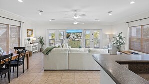 A spacious living area with a large sofa, dining table, and chairs. There is a kitchen area with a countertop and a ceiling fan.