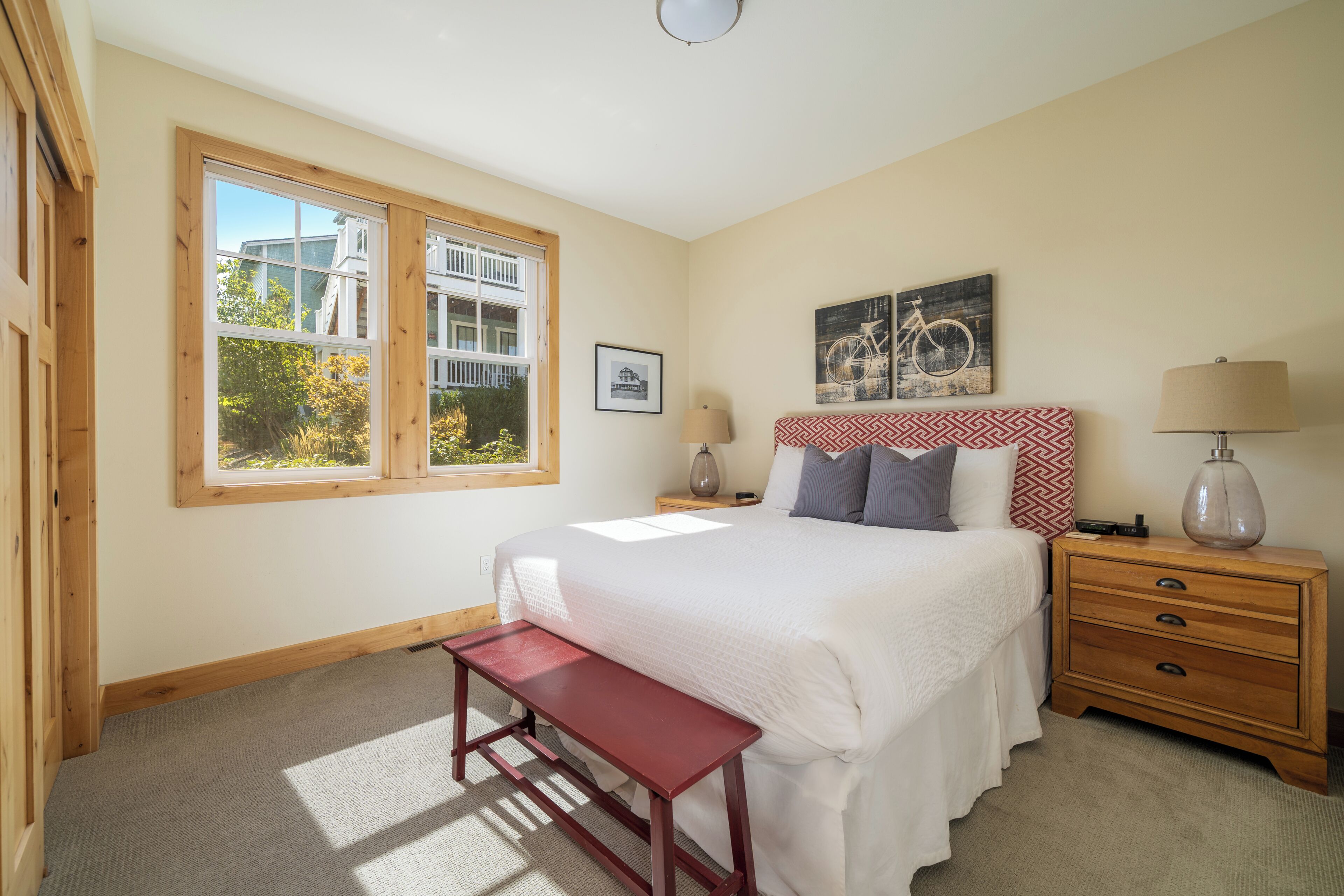 A bedroom with a large window, a bed with white bedding, a wooden nightstand, a bench, and a wall-mounted lamp.