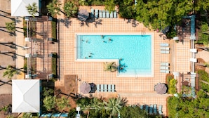 An aerial view of a resort with a central swimming pool, surrounded by lounge areas and palm trees.