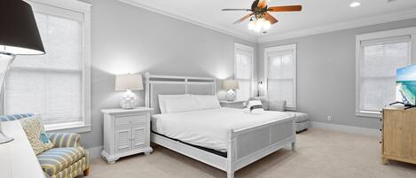 A bedroom with a white bed, two bedside tables, a nightstand, a wooden cabinet, a ceiling fan, and a TV on a wooden stand.