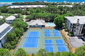 A tennis court surrounded by trees and buildings, with a pool nearby.