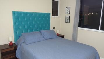 Quarto com cabeceira estofada, cama com roupa de cama azul, mesa de cabeceira com abajur vermelho e janela com vista para a cidade à noite.