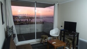 Een zithoek met een bank, een salontafeltje, een witte stoel en een televisie op een dressoir. Een groot raam biedt uitzicht op een stadsbeeld en een wateroppervlak tijdens zonsondergang.