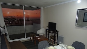 Een eetgedeelte met een tafel voor vier personen, een televisie op een standaard, en een uitzicht op de stad in de schemering door een groot raam.