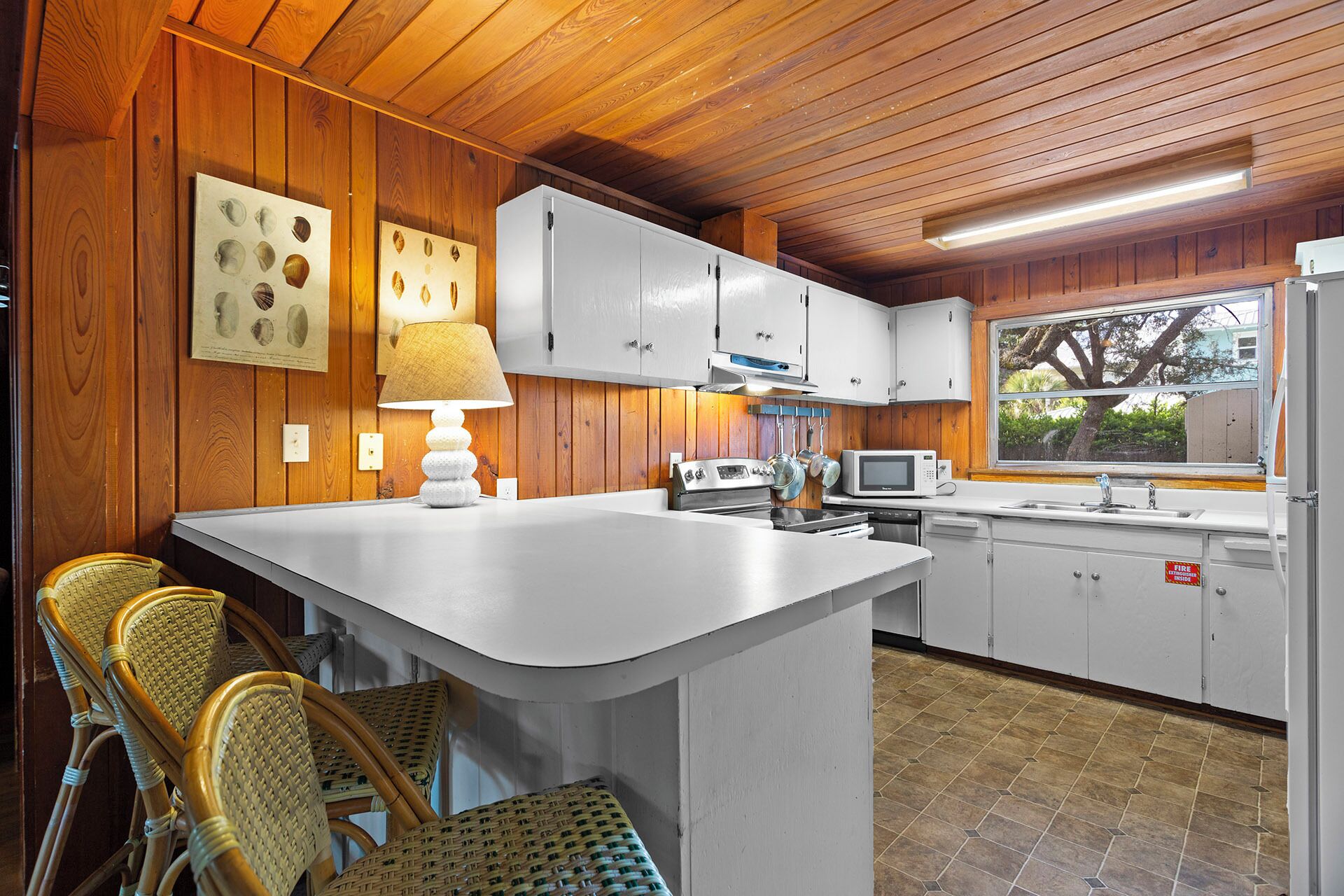 Cocina pequeña con paredes de madera, gabinetes blancos, una isla central, sillas de mimbre y una ventana con vistas.