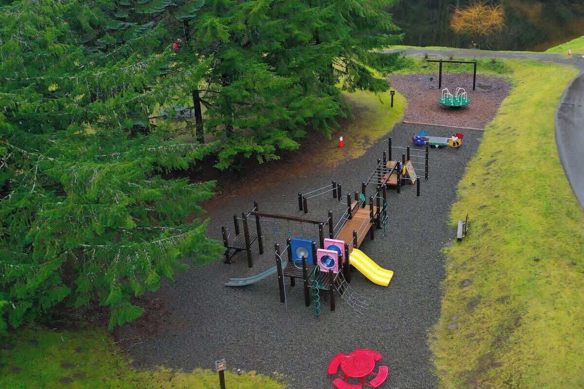 A playground with various play structures, surrounded by trees and a paved path.