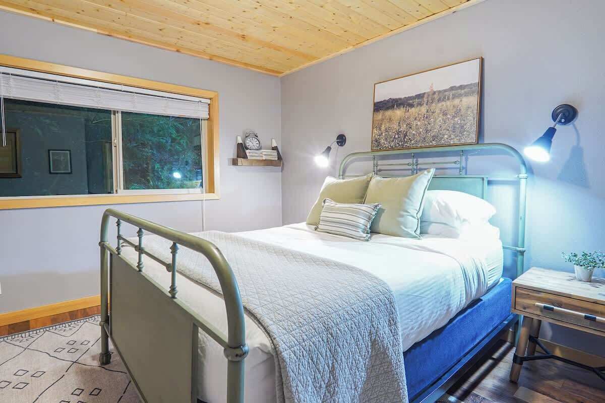 A bedroom with a metal bed frame, a bedside table with a lamp, a window with blinds, and a painting on the wall.