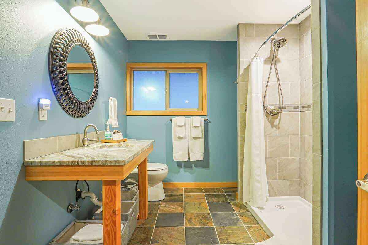 A bathroom with a marble sink, a wooden vanity, a toilet, a shower with a glass door, and a window above the toilet.