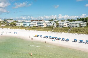 Ein Strandhotel mit mehrstöckigen Gebäuden, einem sandigen Strand mit Sonnenschirmen und blauem Himmel.