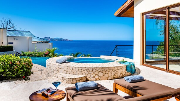 A pool area with lounge chairs and a stone hot tub, overlooking a clear blue sea.