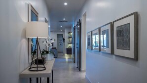 A hallway with framed artwork on the walls, a table with a lamp and orchid, and a door leading to another room. - Steps to the Pool | Private Hot Tub | Red Rock Views | Arcadia #4 (Santa Clara)