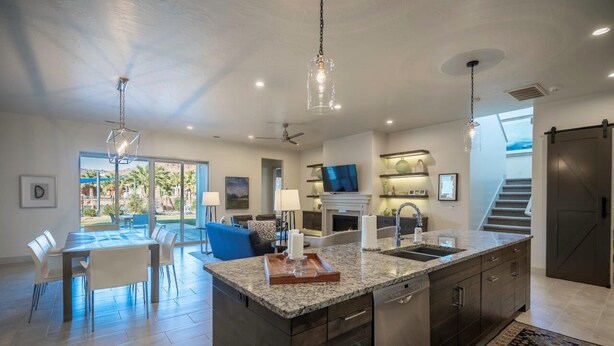 A modern kitchen with granite countertops, dark cabinetry, and a central island. There is a dining area with a table and chairs, a large window with a view, and a staircase leading to an upper level. - Steps to the Pool | Private Hot Tub | Red Rock Views | Arcadia #4 (Santa Clara)