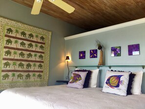 A bedroom with a bed, pillows, a nightstand, a wall hanging, and wall art. - Mampoo Hill-- a Beachfront House 60 feet from the Ocean with optional Jeep (St. John)