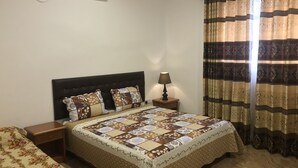 A bedroom with a bed, a nightstand, a lamp, a window with curtains, and an air conditioning unit on the wall.