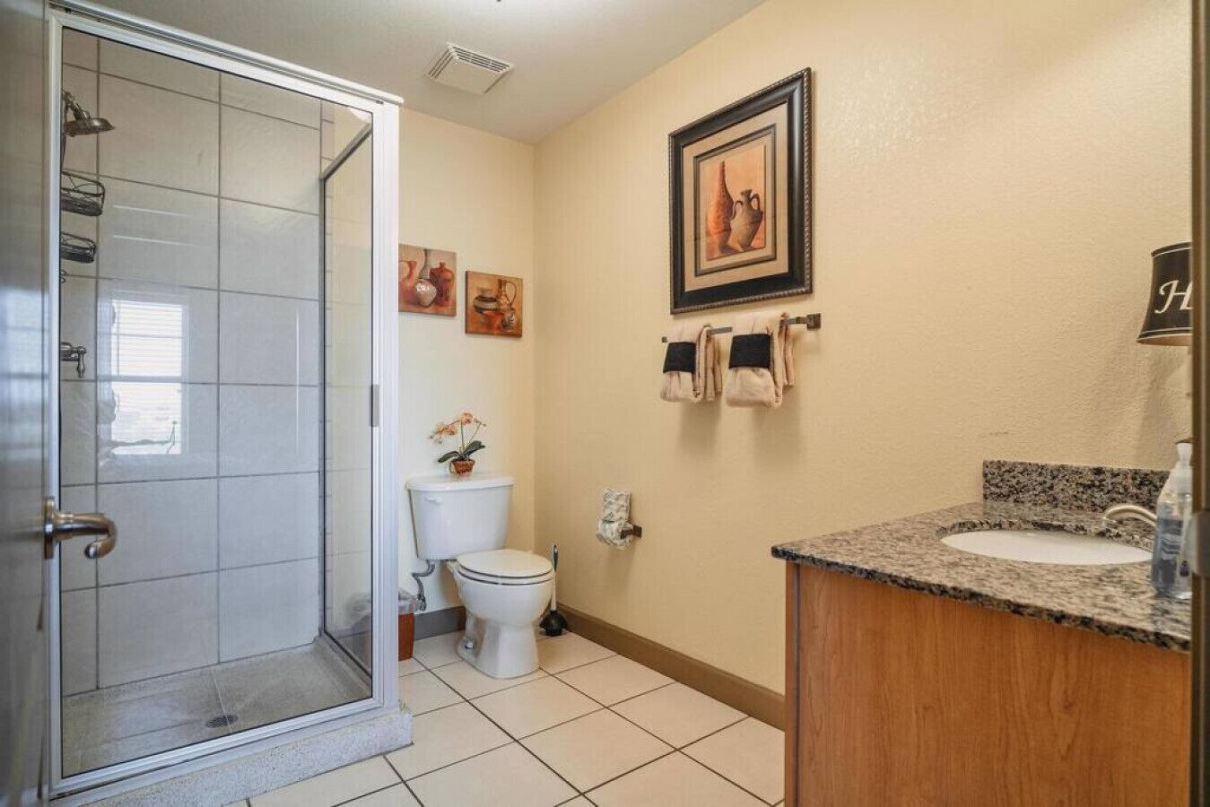 Un baño con cabina de ducha de vidrio, inodoro, lavamanos con encimera de granito y toallas colgadas en un perchero.