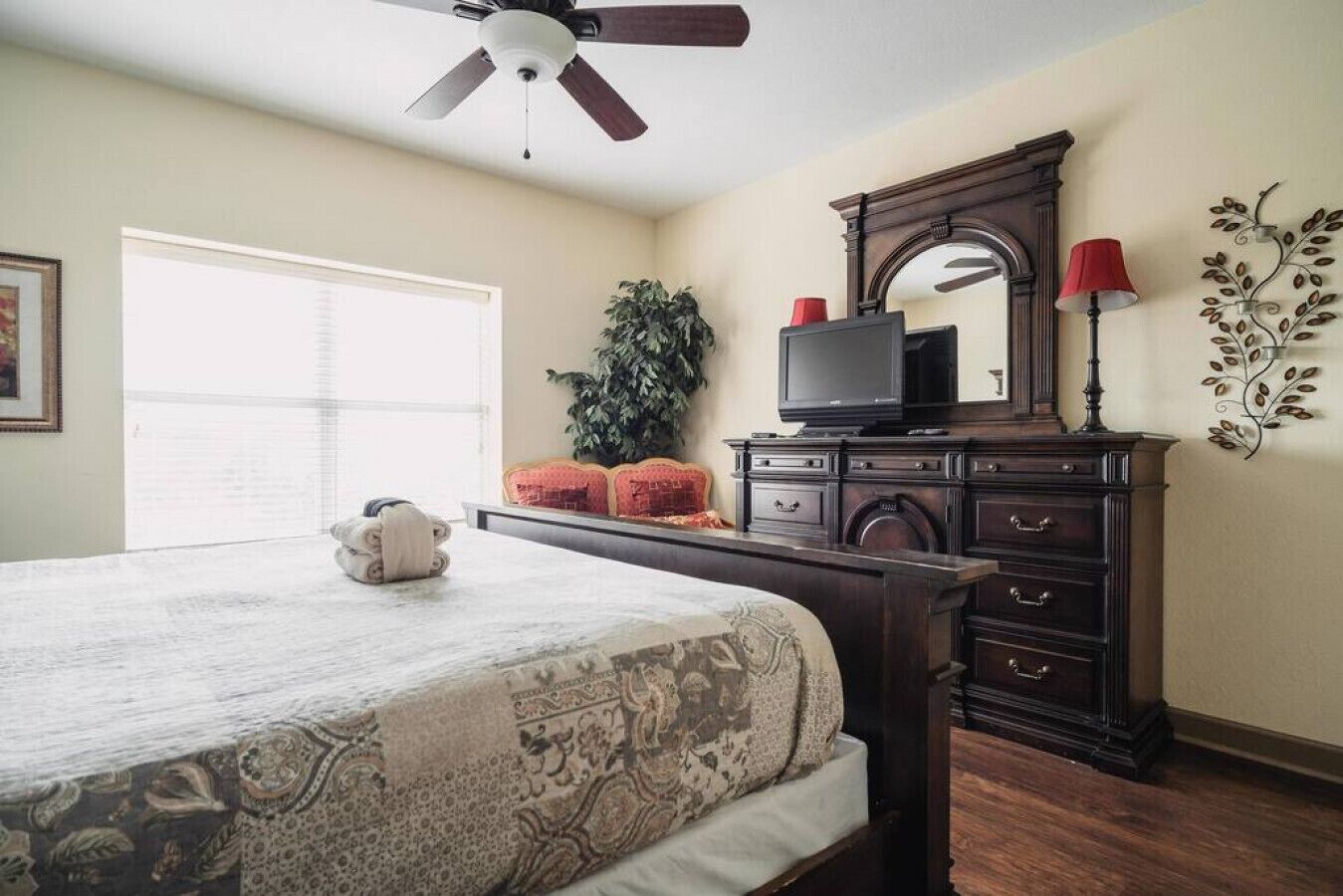 Een slaapkamer met een bed, een dressoir, een televisie, een plant en een raam met jaloezieën.