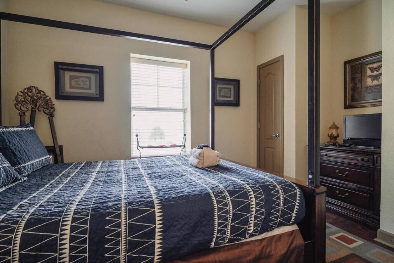 Een slaapkamer met een hemelbed, een raam met zonwering, een deur en een dressoir met een televisie.