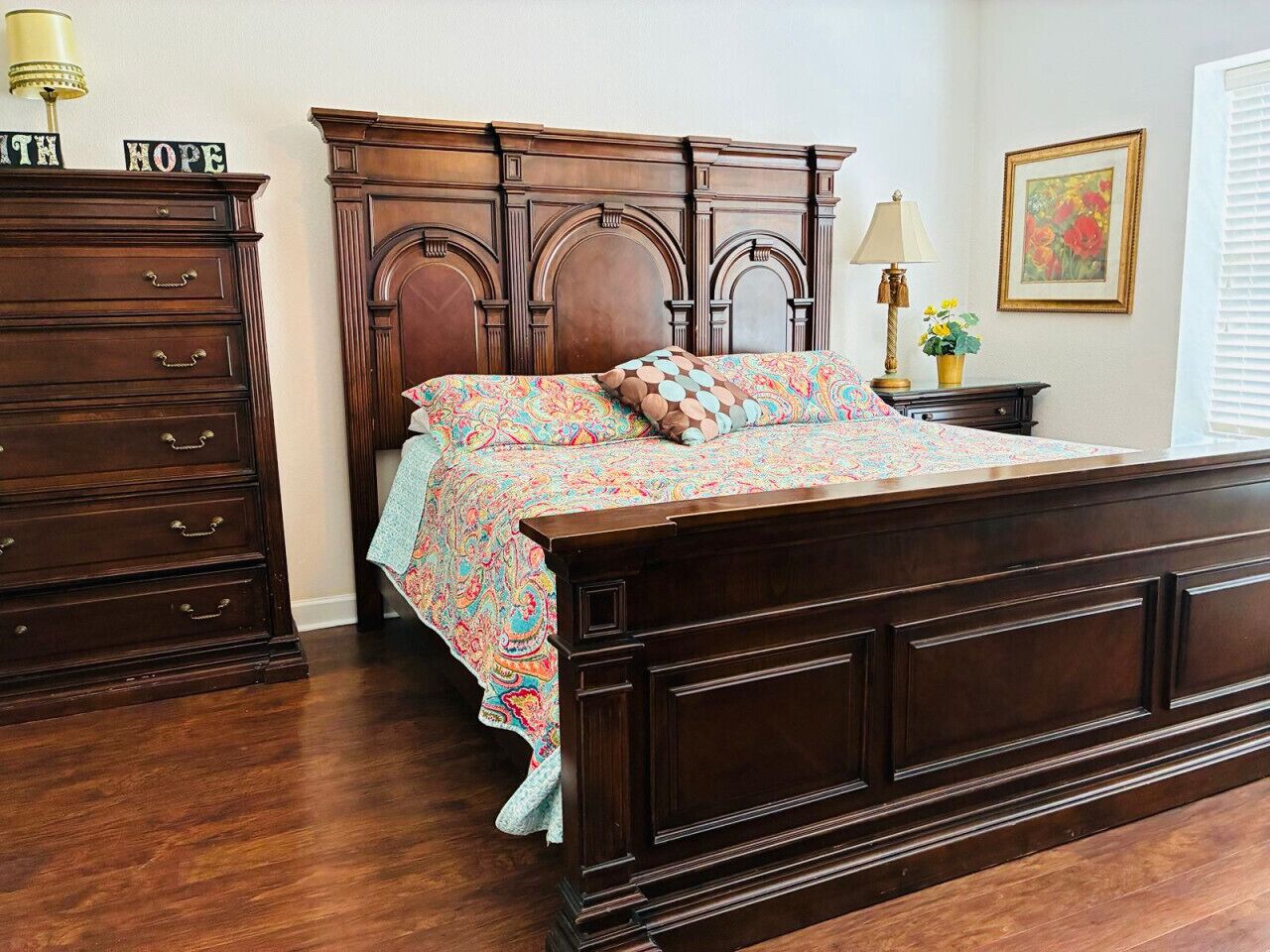 Een slaapkamer met een houten bed, nachtkastje, dressoir en een wand met een ingelijst beeld.