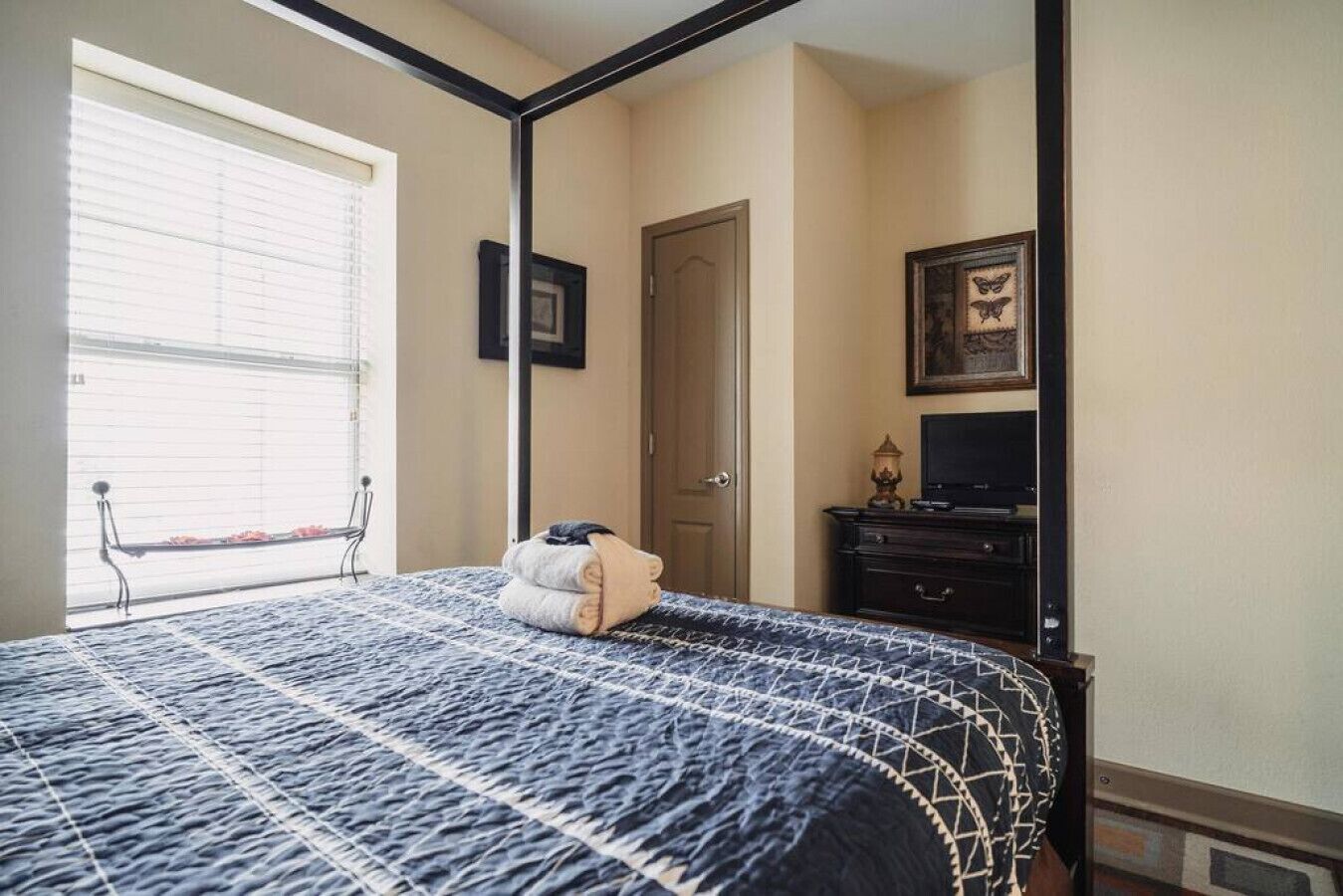 A bedroom with a four-poster bed, a window with blinds, a door, a dresser, and a framed picture on the wall.