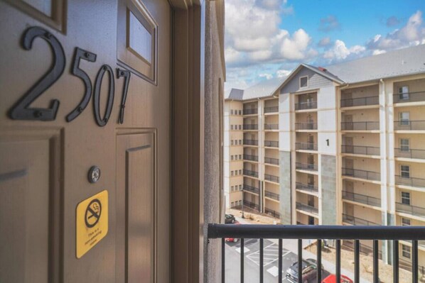 Un balcón con vista a un edificio de varios pisos, una puerta con el número 2507 y un cartel de prohibido fumar.