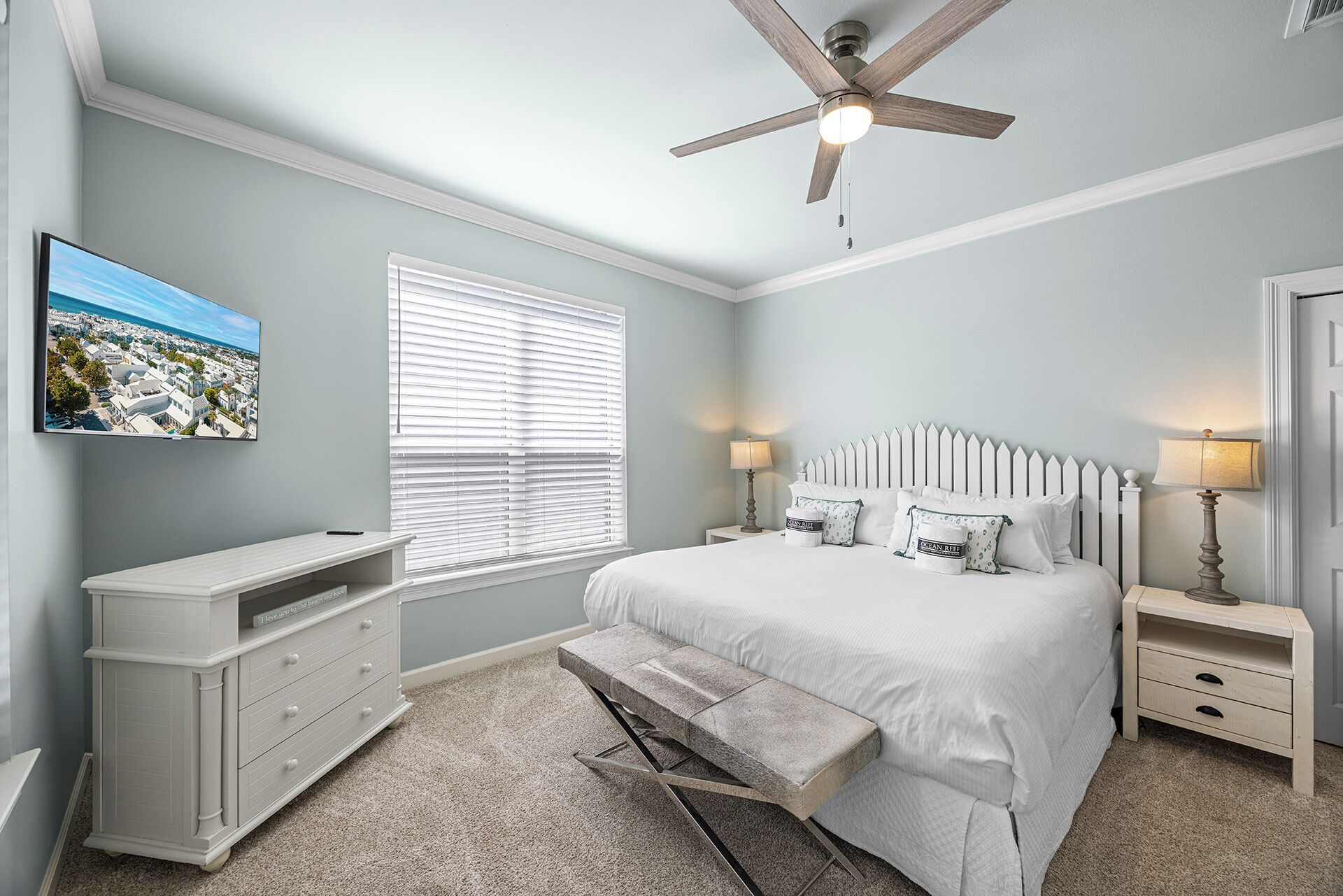 A bedroom with a bed, a nightstand, a bench, a ceiling fan, a wall-mounted TV, and a window with blinds.